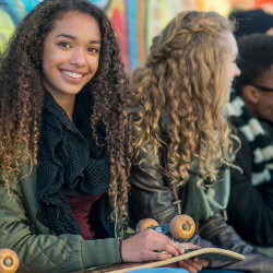 Teenager smiling while sitting against a spray painted wall with a skateboard on her lap.