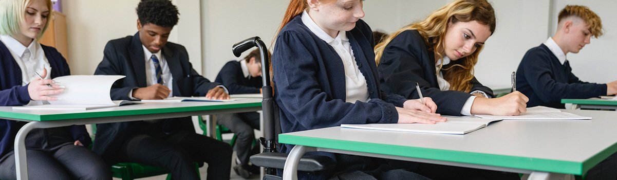 Une adolescente en fauteuil roulant avec ses camarades de classe écrivant leur examen dans des cahiers de composition.
